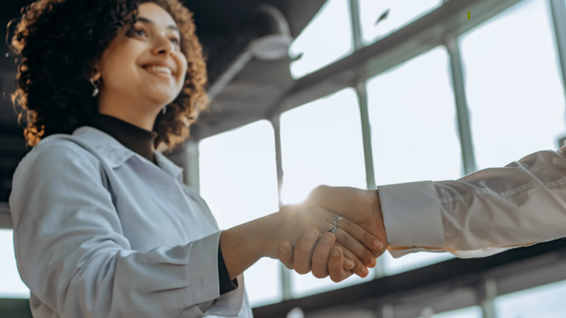 A smiling professional shakes hands with another person in a bright, modern indoor setting, suggesting a business agreement or partnership.
