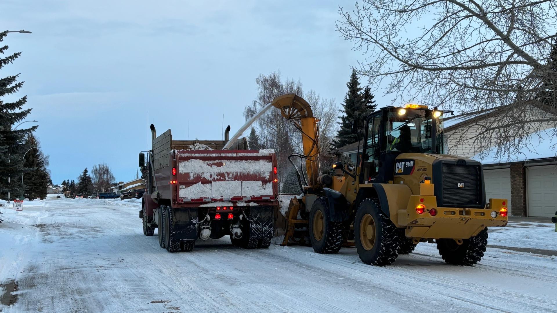 A municipal snow removal crew uses a front-end loader to blow snow into a dump truck along a residential street during winter conditions.