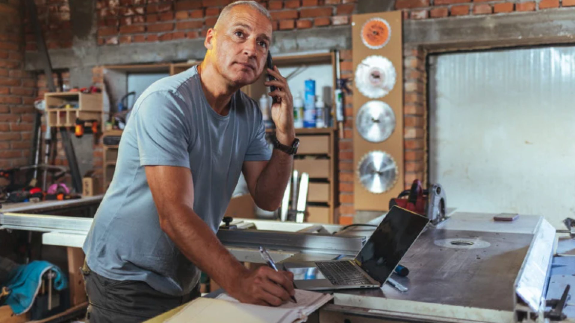 A man in a workshop talks on the phone while taking notes beside tools and equipment, suggesting he is managing or starting a business.
