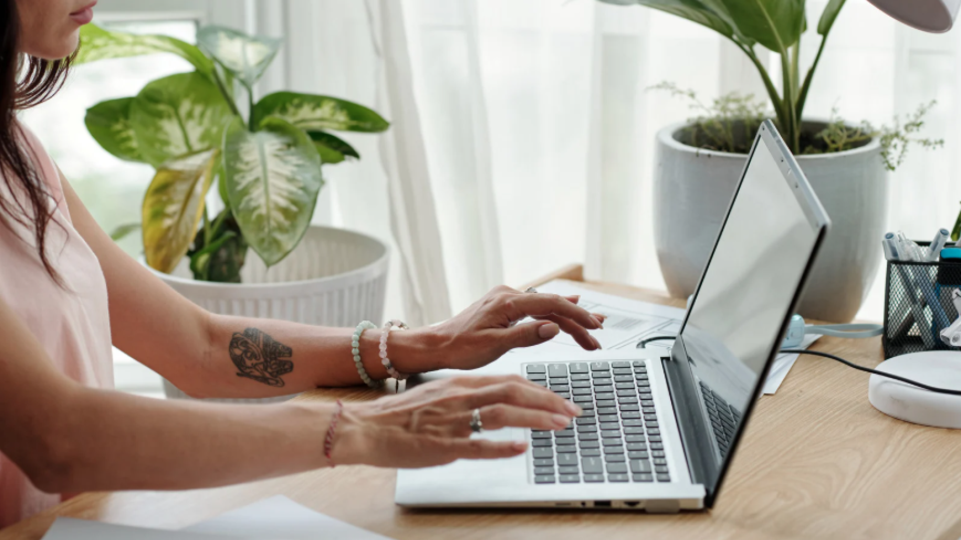 A person works on a laptop at a home desk with plants nearby, suggesting a home-based or remote business setup.