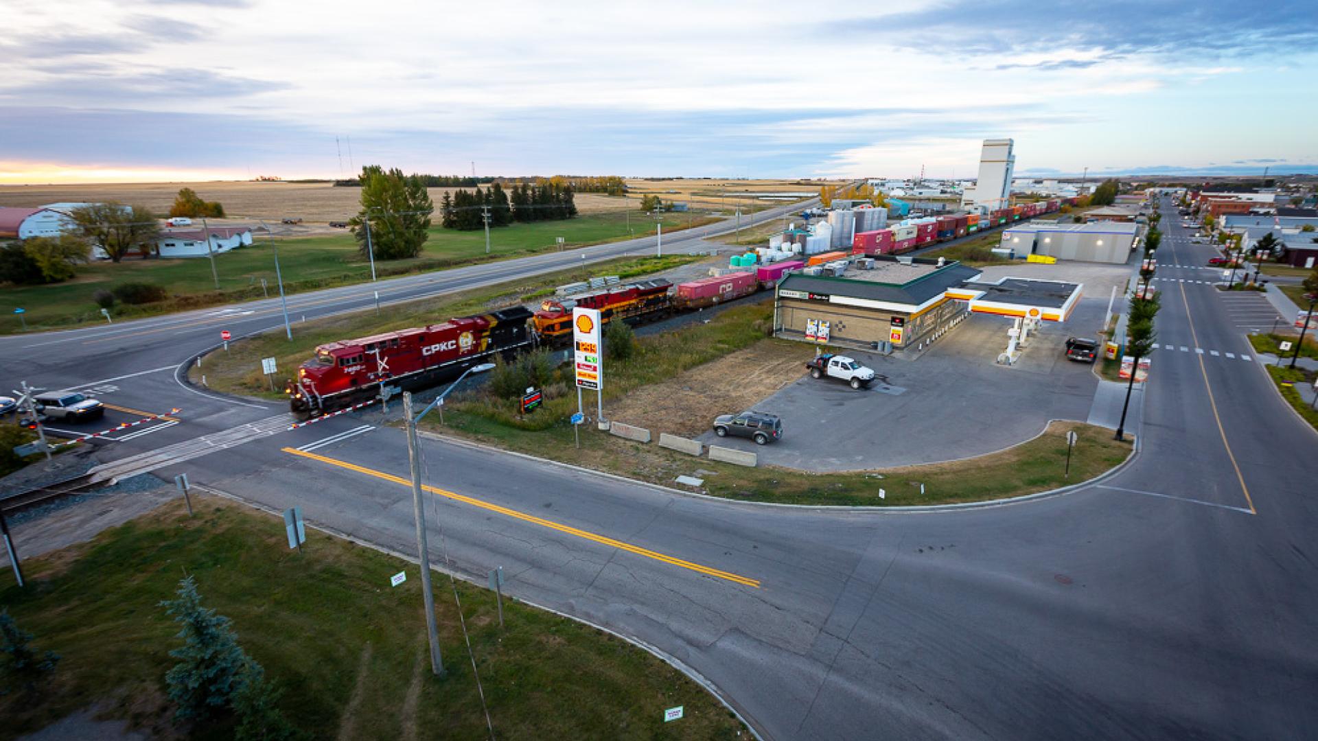An aerial view of a freight train crossing a road beside a gas station in Crossfield, with nearby businesses, rail yards, open farmland stretching into the distance.