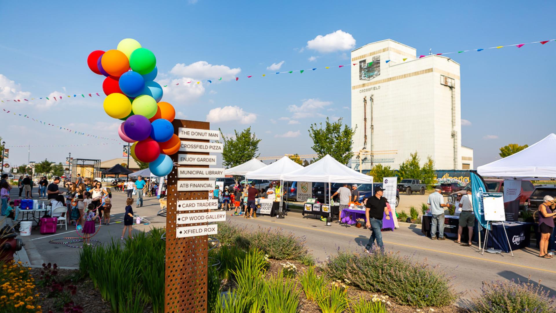 A lively street festival in Crossfield with vendor tents, families, and colourful balloons, set against the town’s grain elevator on a sunny day.