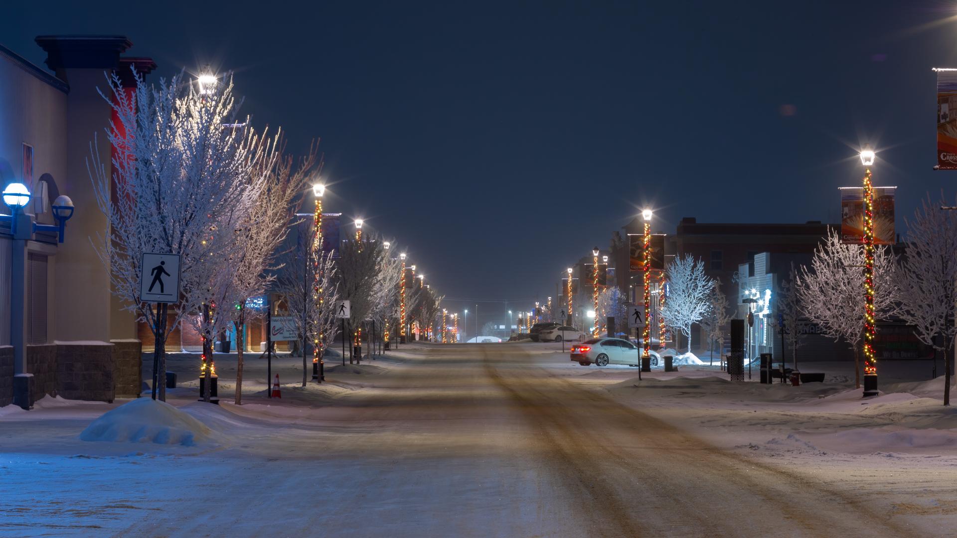 Snow-covered Railway Street in Crossfield at night, looking south, with frosted trees and streetlights wrapped in holiday lights lining both sides of the quiet main street as it stretches into the distance.